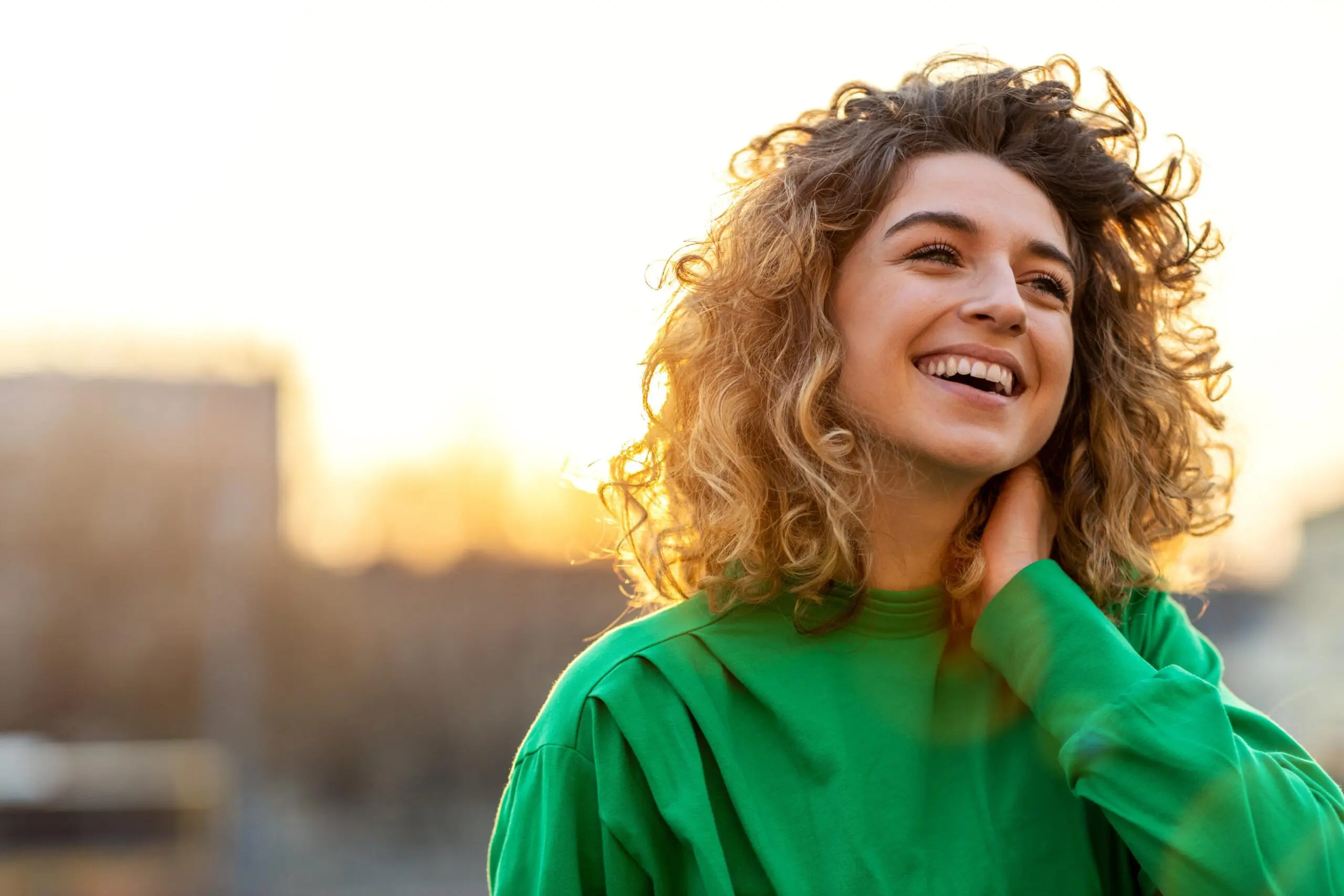 Young woman with curly hair smiling outdoors during sunset.