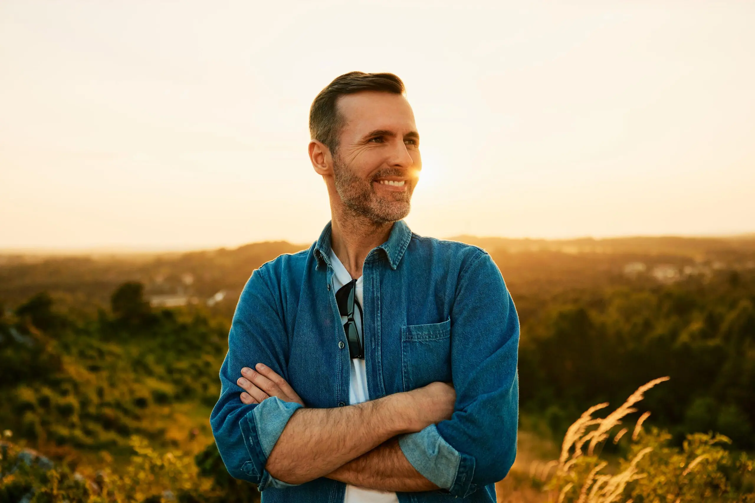 Man in a denim shirt standing outdoors at sunset, smiling.